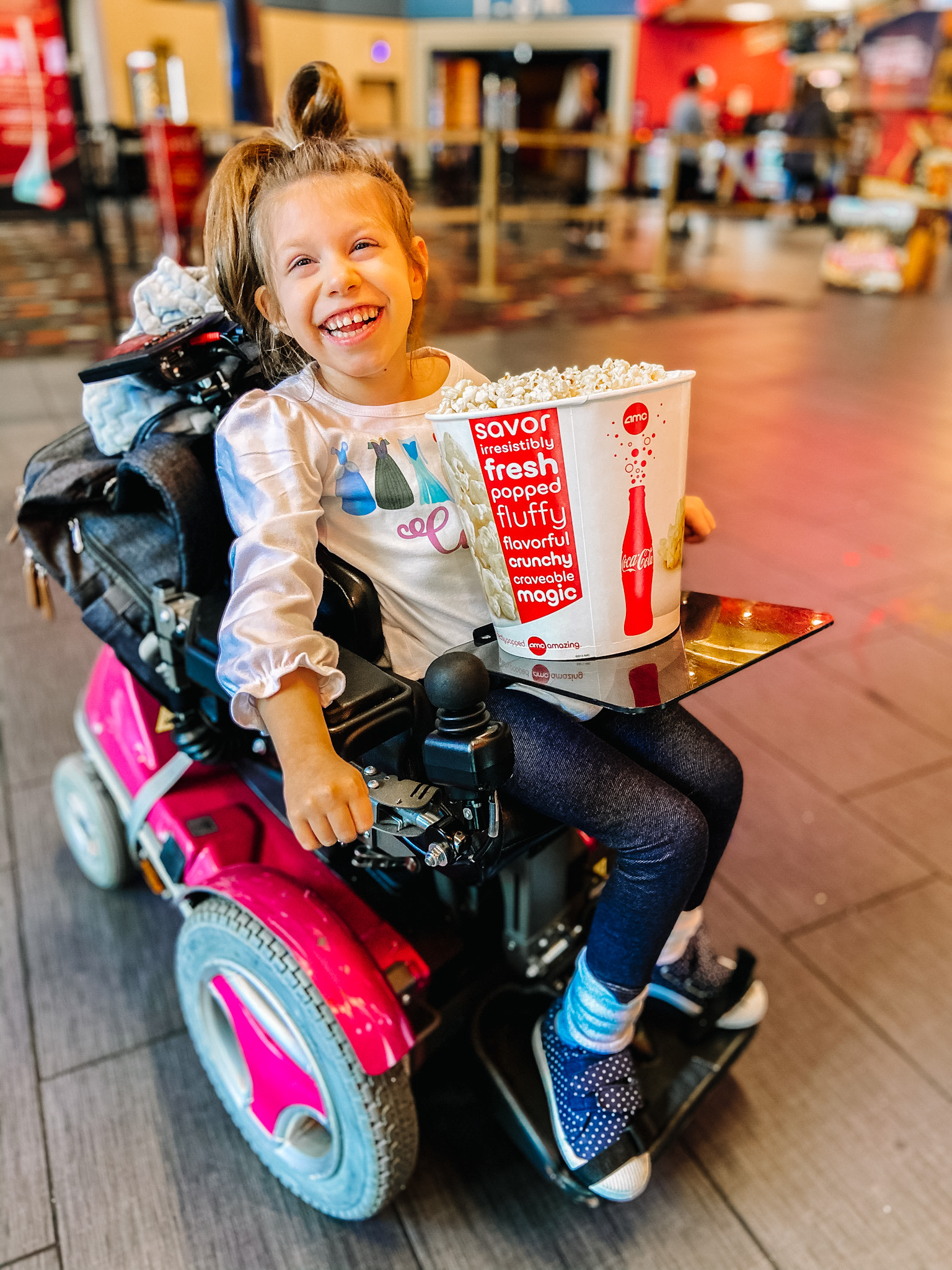 A young girl with a joyful smile sits in a bright pink power wheelchair, holding a large bucket of popcorn at a movie theater. She wears a white long-sleeve shirt with a colorful design, dark leggings, and polka-dot shoes. The theater lobby is visible in the background.